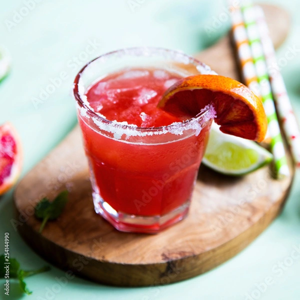 Fototapeta Blood orange and lime margarita cocktail with slices of fresh fruit and ice on optimistic green background.