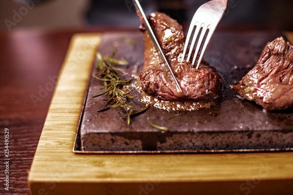 Fototapeta sirloin steak on a very hot stone being cooked by a man to his own taste on a wooden table with a knife and fork