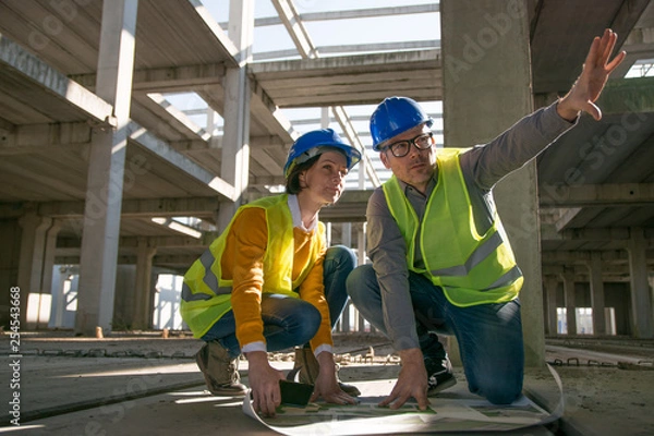Fototapeta Two architects kneeling on the floor and discussing over blueprint
