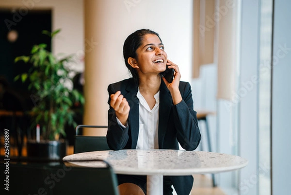 Fototapeta An elegant Indian woman sitting inside a cafeteria. She is smiling while talking to a friend over the phone. 