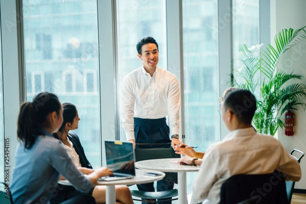 Fototapeta A gorgeous, certain and fit Asian Chinese man seats a gathering with his group amid the day in the workplace. He is expertly wearing a shirt and pants and is motioning as he talks.