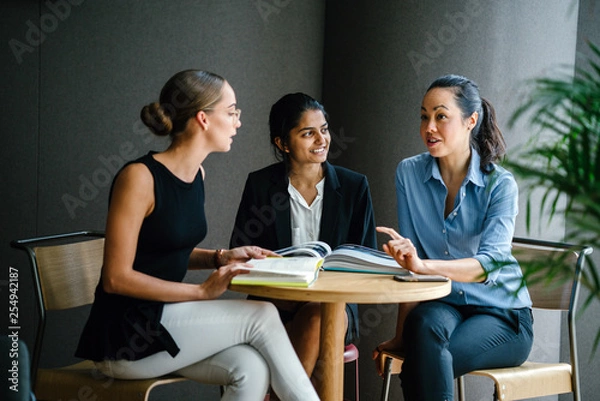 Fototapeta Portrait of a diverse group of three professionally dressed young woman (Chinese, Indian and Caucasian) having a casual discussion around a table in a meeting room during the day.