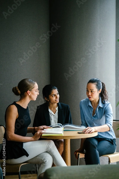 Fototapeta A young Caucasian woman is having a casual business meeting with her team in a meeting room. They are sitting and having an animated conversation. The team is diverse (Indian and Chinese). 