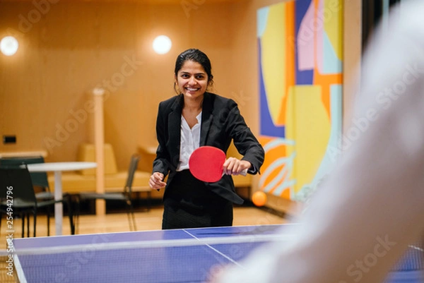 Fototapeta A young and attractive Indian Asian woman in a suit playing table tennis with her colleague in the office during a break. She's having lots of fun. Image taken with a blurred art work as background.