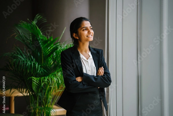 Obraz Portrait of a young Asian Indian professional woman in a meeting room standing by the glass window, smiling with her arms crossed. She looks optimistic, happy and confident