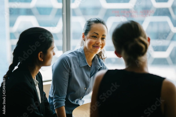 Fototapeta Image of a young Chinese Asian woman manager chatting with her team in their office. She smiles back as she listens to her colleague talking about their new project.