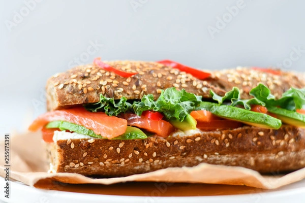Fototapeta sandwich from a cereal baguette with avocado, salmon, cream cheese, tomatoes and lettuce leaves on a white plate.  light background, selective focus and copy space.