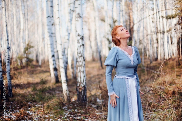 Obraz Beautiful young girl of European appearance with red hair and blue eyes on the background of spring birch forest. Happy woman  looking at the sky in nature. Illustration to a book or a fairy tale.