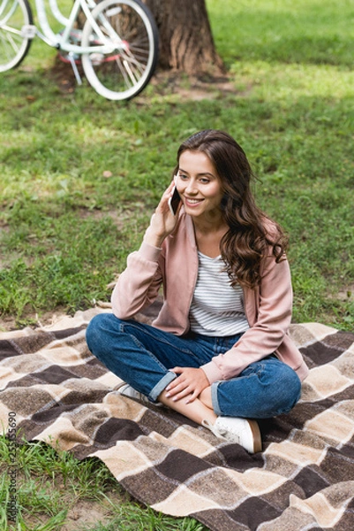 Fototapeta cheerful girl talking on smartphone while sitting on checkered blanket in park