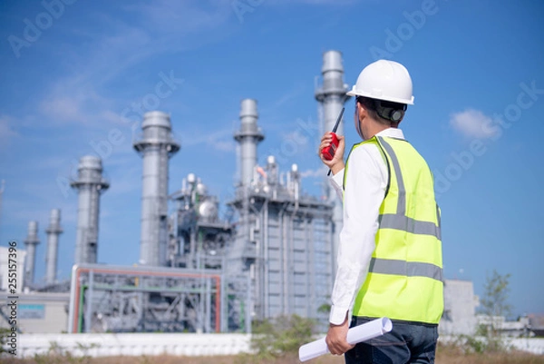 Fototapeta Industrial engineer standing in front of a large  Power plant with safety helmet with gas turbine industry plant background