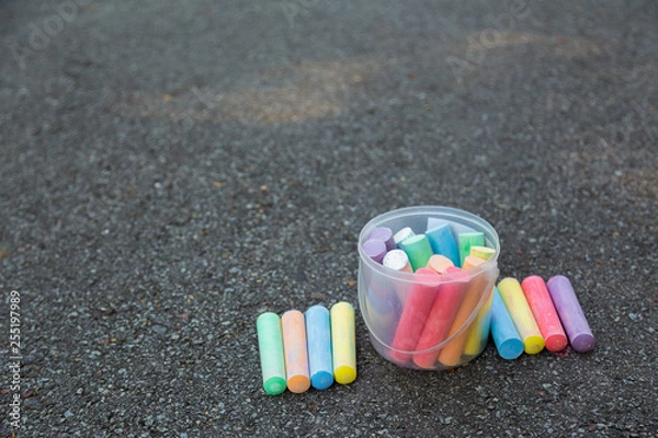Fototapeta Plastic transparent box full of new colorful chalks outdoors isolated on surface of grey grunge pavement as background. Horizontal color photography.