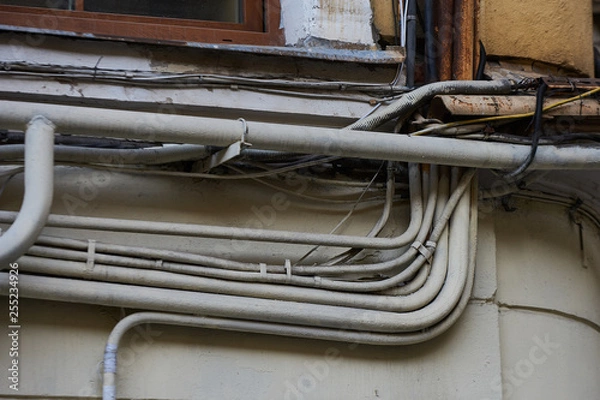 Fototapeta Wires - electrical, telephone, antenna, computer, network on the wall of a dilapidated house with peeling plaster, separate wires in metal pipes with protection