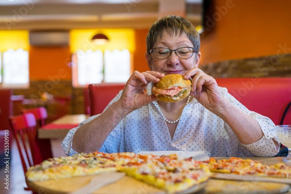 Fototapeta Aged business woman in a cafe-restaurant is glad that she eats fast food. The problem with overweight and junk food does not interest her.