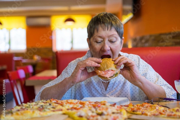 Fototapeta An elderly lady works a lot and decided to have a snack in a cafe restaurant. Fast food is not good for health. Overweight and fatigue.