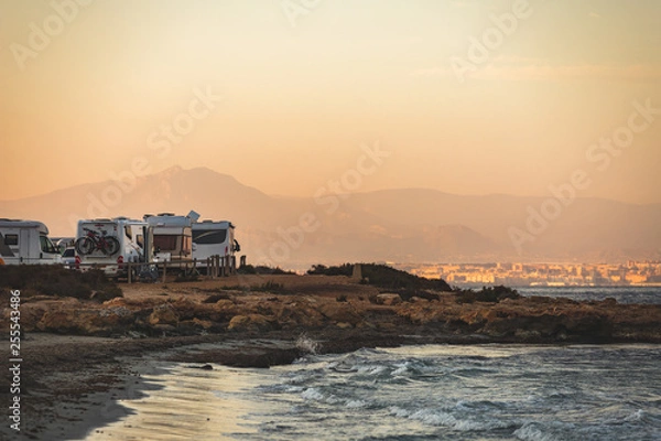 Fototapeta Motorhome making wild camp on the beach at a beautiful sunset or sunrise with mountains in the background. Caravan parked on the beach in front of the blue sea in a beautiful place of wild nature