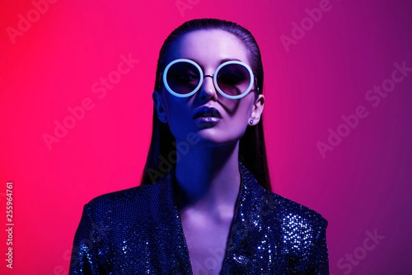 Fototapeta Moda dziewczyna z długimi włosami i okrągłe okulary przeciwsłoneczne w czarnej lśniącej sukience pozuje w świetle neonu w studio