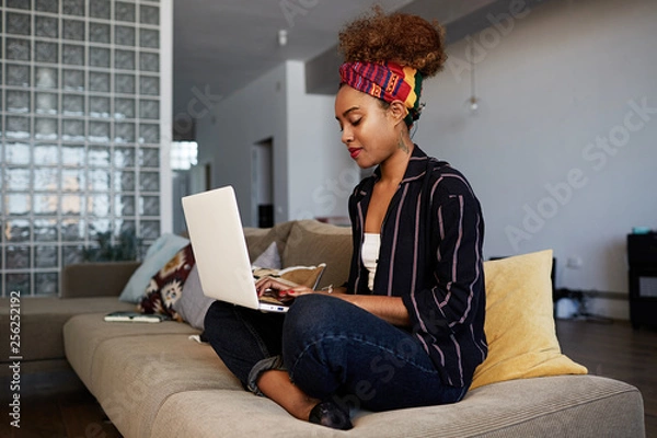 Fototapeta Young american african female blogger working remotely on digital netbook with internet text. African woman copywriter typing article for website on keyboard of modern laptop computer at home.