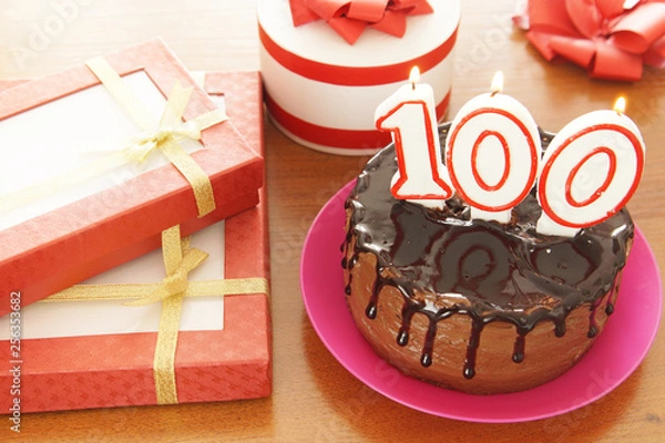 Fototapeta Birthday celebration at one hundred years. A cake with burning candles in the form of numbers and a lot of gifts on a table