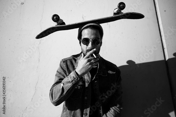 Obraz man posing on a urban wall smoking and wearing sunglasses with a skate on his head