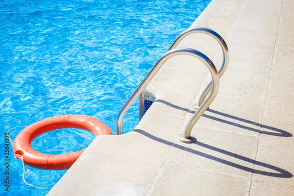 Obraz Closeup view of the swimming pool edge with staircase and a red ring life buoy floating on the blue water during a sunny summer day