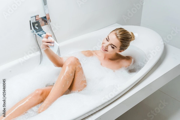 Fototapeta attractive and smiling woman taking bath with foam and taking selfie in bathroom