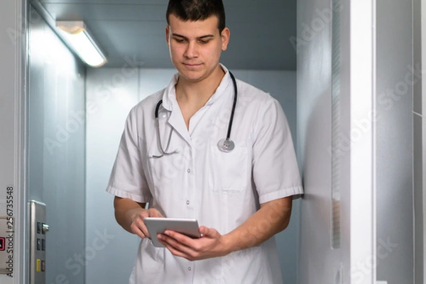 Fototapeta Bright photo of male doctor in uniform with stethoscope coming out of the elevator and using computer tablet in hospital. Modern medical concept