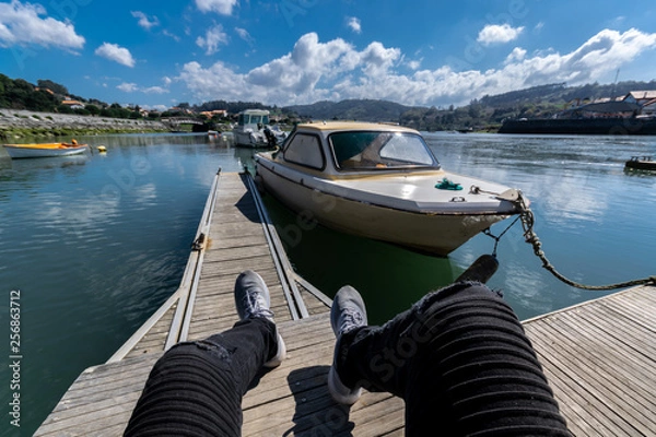 Obraz persona sentada en un puerto con un lago o rió de fondo y una barca evitada para no ser reconocida cielo despejado