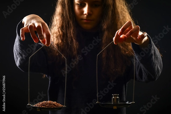 Fototapeta Emotional portrait of a girl with long curly hair weighing grain and weights on scales against a black background