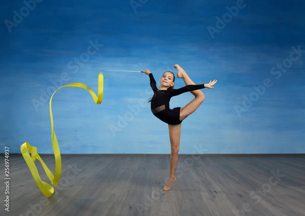Obraz Girl gymnast in a black gymnastic swimsuit does a wave in the air with a yellow ribbon on a blue sky background.