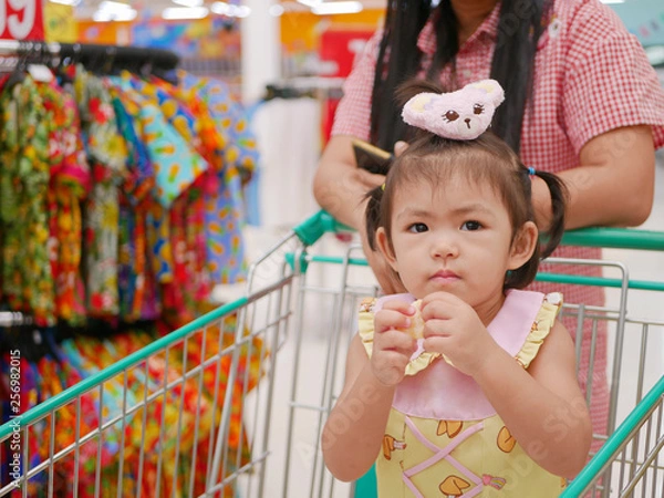 Fototapeta Little Asian baby girl, 24 months old, eating snack and standing in a shopping cart while her mother does a shopping in a supermarket / mall