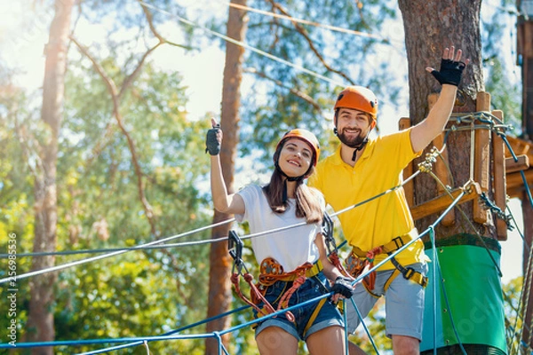 Fototapeta Young woman and man in protective gear are standing on rope bridge hanging on high trees, posing and smiling. Rope park with obstacles and ziplines. Extreme rest and summer activities concept.
