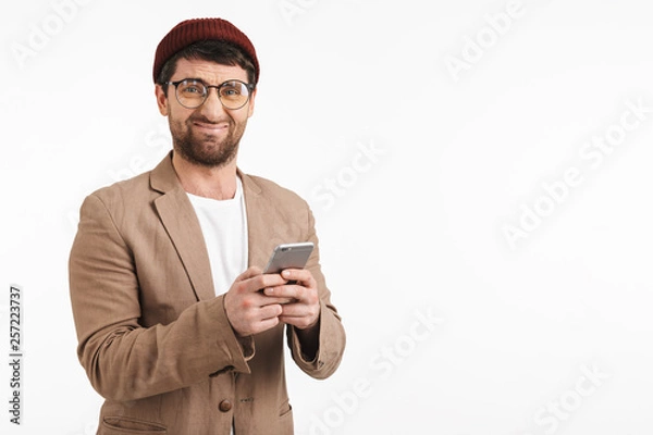 Fototapeta Photo of caucasian man 30s wearing hipster hat smiling while holding and using smartphone