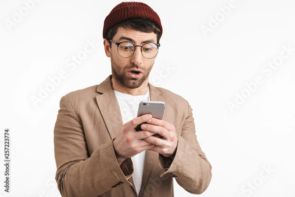 Fototapeta Photo of excited man 30s wearing hipster hat smiling while holding and using smartphone