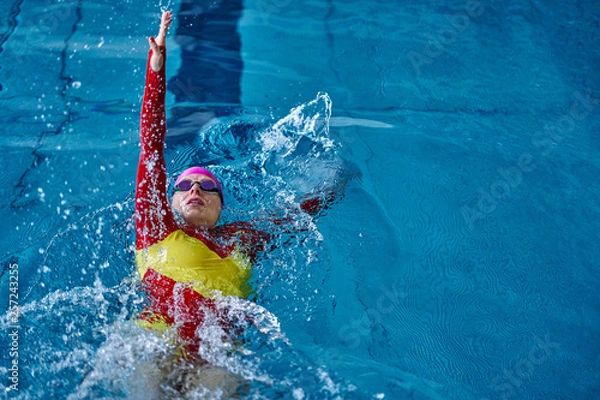 Fototapeta Female athlete in a red-yellow swimsuit is swimming on his back. Splashes of water scatter in different directions.