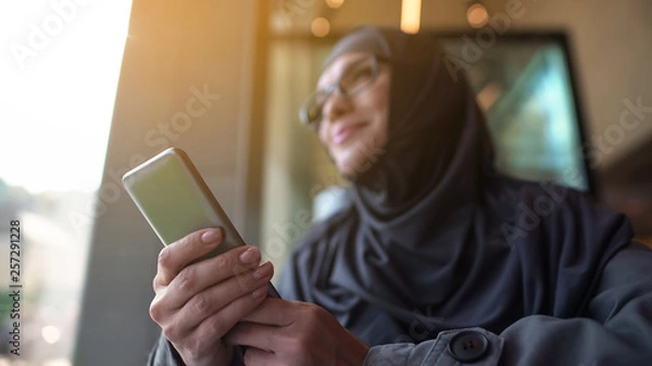 Fototapeta Cheerful Muslim lady holding phone in hand, looking outside through cafe window