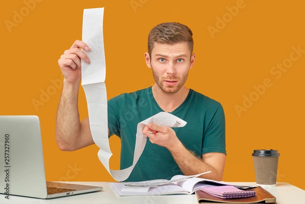 Fototapeta The indignant man holds checks, not understand from where the big sums, not ready to unforeseen expenses. Isolated on an orange background in the Studio