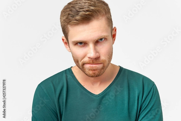 Fototapeta Puzzled man keeps lips pressed, has frustrated facial expression, frowns face in displeasure, being deep in thoughts. Male dressed in casual outfit, models over white wall
