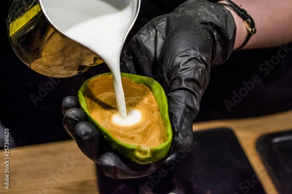 Fototapeta The barista in black gloves prepares a cappuccino with latte an art in avocado. Unusual cup. Fresh and beautiful coffee with milk.