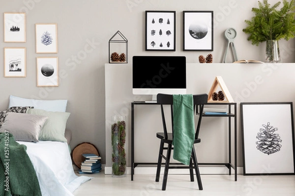 Fototapeta Green cloth on black wooden chair at desk with computer, wooden triangle and cones, real photo with gallery of posters on empty grey wall