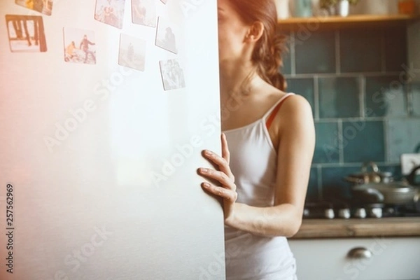 Obraz A young woman is looking into an open fridge while holding the door with her left hand. Diet concept.