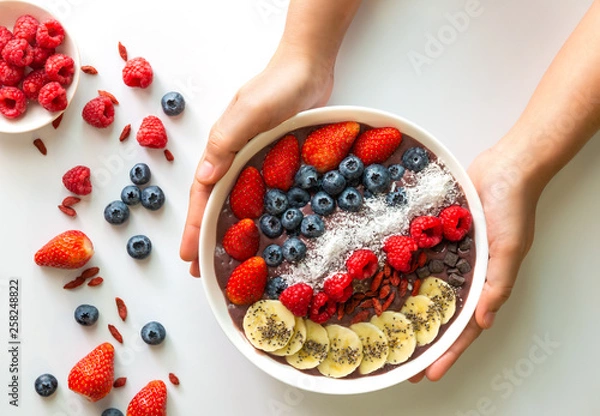 Fototapeta Woman hands holding an Acai Berry superfoods smoothies white bowl with chia seeds, strawberry, goji berry, coconut, blueberry, raspberry toppings, and chocolate chip on white background