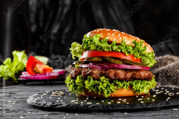 Fototapeta Beef burger with tomatoes, red onions, cucumber and lettuce on black slate over dark background. Unhealthy food