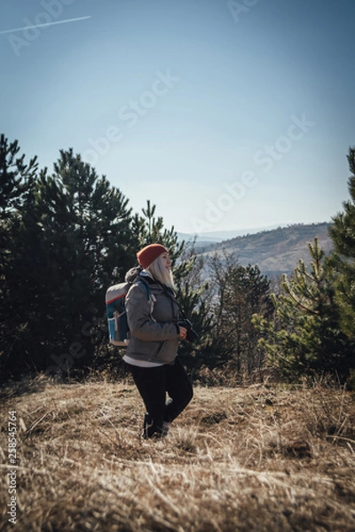 Obraz Hiking girl with backpack standing on the meadow with pine forest on the mountain and observing locality.