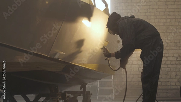 Fototapeta In slow motion a professional man (guy) in a garage (service) in working clothes dressed in a sweater sets up a paint gun (spray gun) and starts painting (putting a layer of ceramics) a yacht.