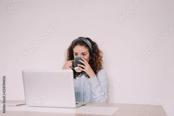 Fototapeta A young woman is sitting in front of her computer while drinking a cup of coffee. Look at something pretentiously on the screen of your laptop. 