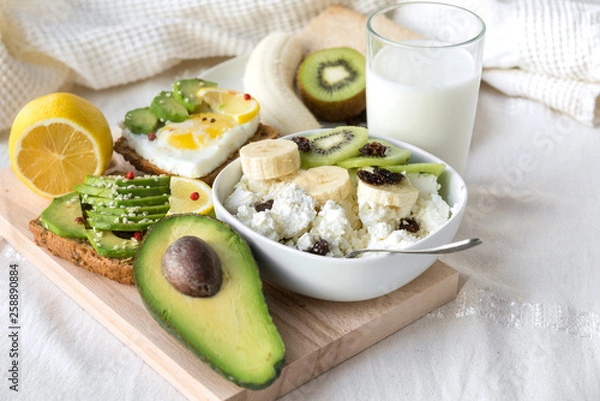Fototapeta cottage cheese with banana slices ,kiwi and raisins, half avocado, avocado sandwich, lemon, sesame, egg,  glass of milk on a white tablecloth
