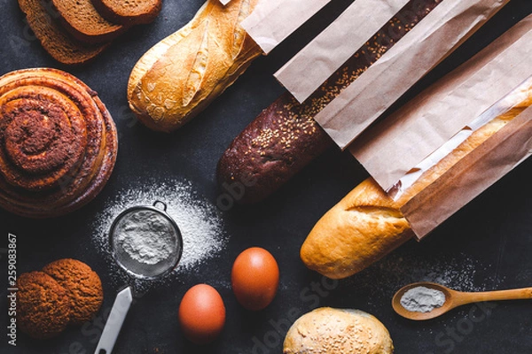 Fototapeta Baking ingredients for flour and rye bakery products. Fresh crisp bread, baguette packaged in paper bag and buns on a black background.