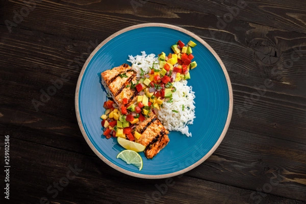 Obraz Delicious grilled salmon steak on a blue plate with jasmine rice and sauce made of mango, bell pepper, onion and avocado. Top notch food. Top view shot above.