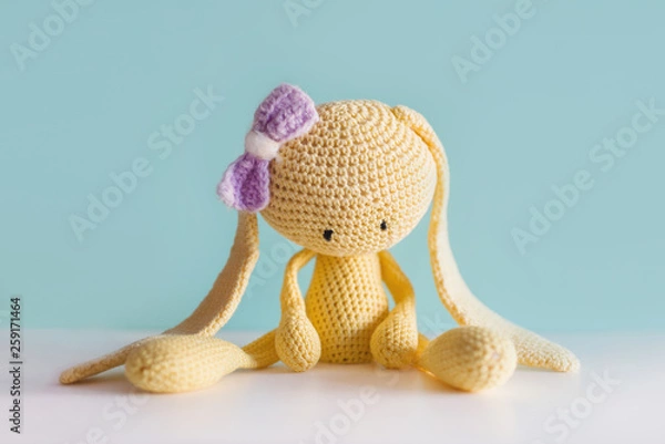 Fototapeta Soft toy knitted hare. Sitting yellow sad plush hare with a violet bow. Amigurumi.