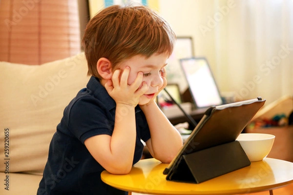 Fototapeta Child using tablet pc on bed at home. Cute boy on sofa is watching cartoon, playing games and learning from laptop. Education, fun, leisure, happiness, modern computer technology and communication.